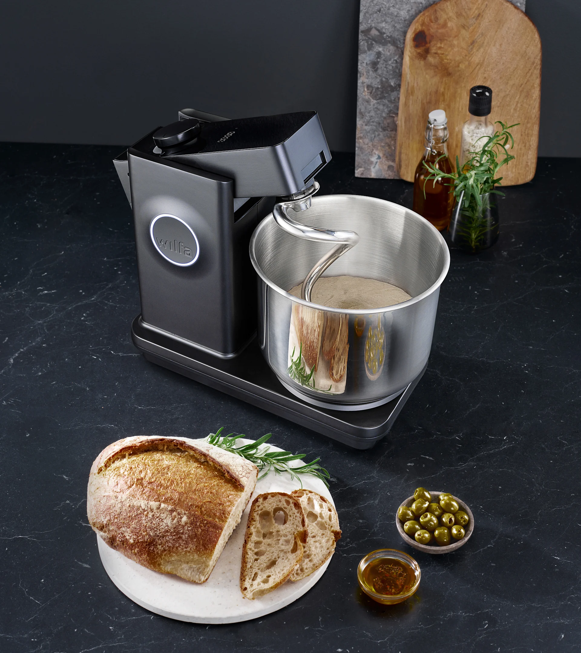 Schwarze Küchenmaschine knetet Teig in einer Edelstahlschüssel, daneben rustikales Brot, geschnittenes Brot, Oliven und Olivenöl auf dunkler Arbeitsplatte.