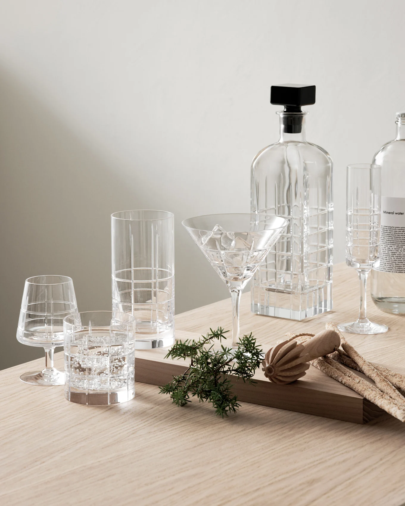 Klare Glaskaraffe, verschiedene gemusterte Gläser und eine Mineralwasserflasche mit Wacholder und Grissini auf Holztisch.