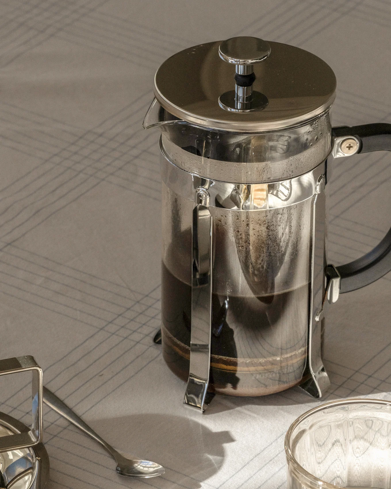 Eine French Press Kaffeemaschine mit dunklem Kaffee auf einer gestreiften Tischdecke, neben einem Löffel und Glas.