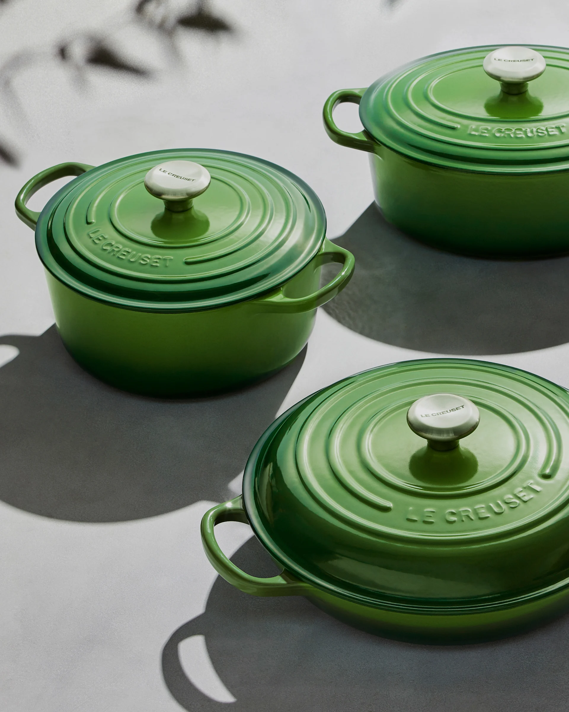 Drei grüne Le Creuset Töpfe mit Deckeln und silbernen Knöpfen auf hellem Untergrund mit Schatten.