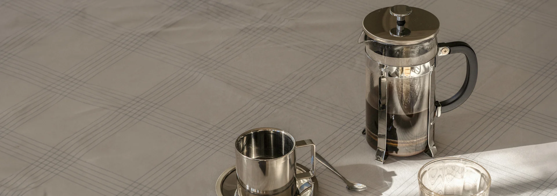French Press mit Kaffee, eine Metalltasse mit Untertasse und Löffel auf einem hellen, karierten Tischtuch im Sonnenlicht.