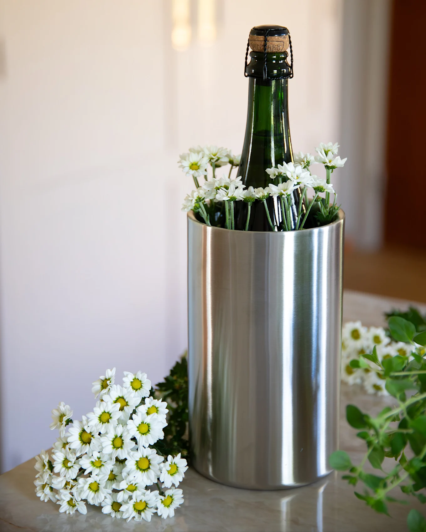 Eine dunkelgrüne Flasche mit Korken in einem Edelstahlkühler, umgeben von weißen Gänseblümchen auf einer hellen Marmorfläche.