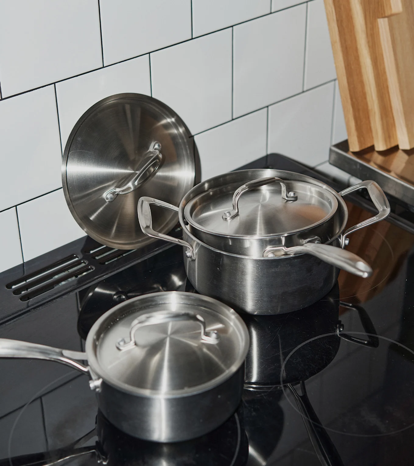 Töpfe und Deckel aus Edelstahl auf einem schwarzen Induktionskochfeld mit weißem Fliesenspiegel und Holzschneidebrettern.
